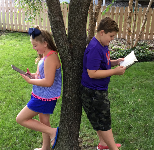 Two children leaning against opposite sides of a tree. The girl is reading on a tablet computer; the boy is reader a traditional print book.