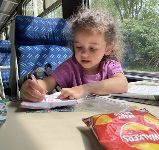 A preschooler drawing in a notebook while riding a train.