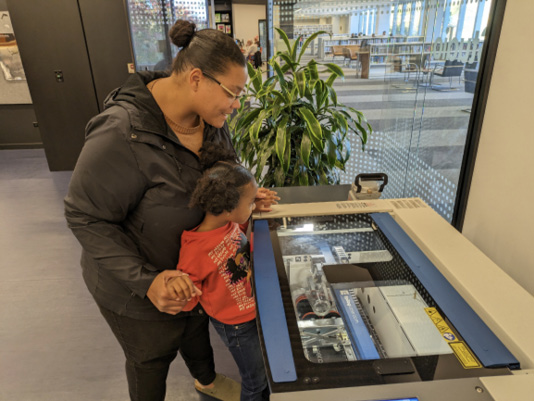 A child and her mom experiment with a laser cutter at Skokie (IL) Public Library.