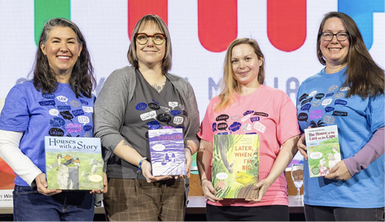 Members of the Batchelder committee displaying their award and honor books and modeling their awesome and apropos t-shirts featuring speech bubbles that contain the word read in multiple languages.